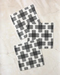 Three square Cross Hatch napkins by Rosa Kichline, made from recycled materials and featuring a black intersecting line pattern, are arranged diagonally with slight overlap on a light marble surface for a modern, geometric effect.