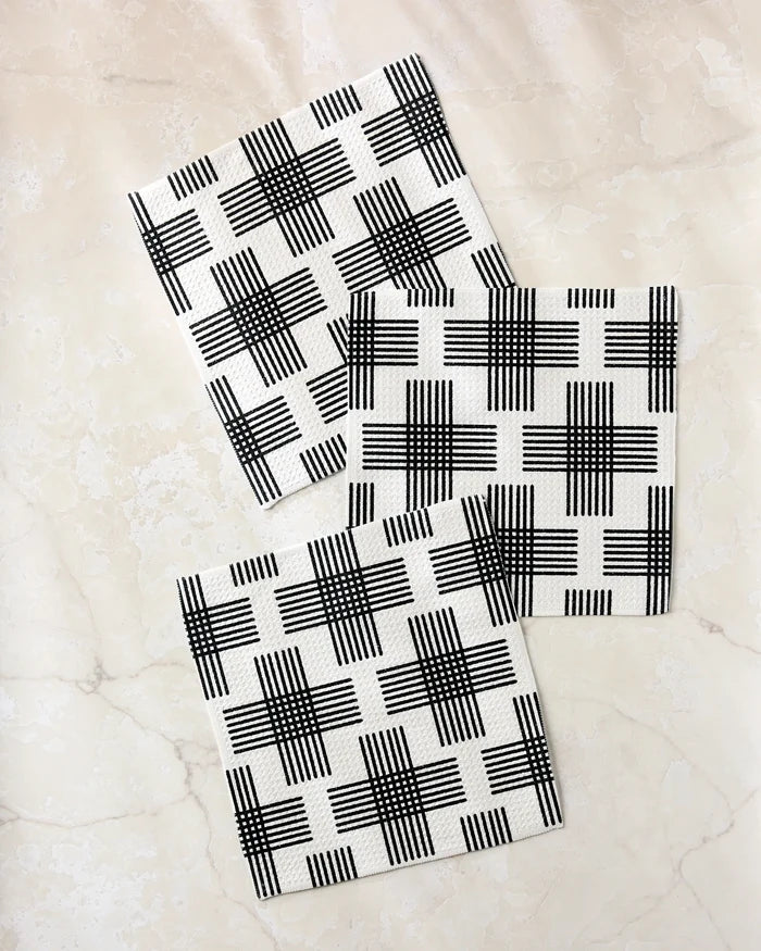 Three square Cross Hatch napkins by Rosa Kichline, made from recycled materials and featuring a black intersecting line pattern, are arranged diagonally with slight overlap on a light marble surface for a modern, geometric effect.