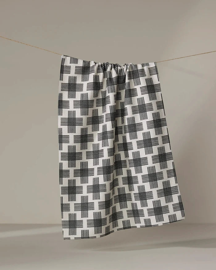 The Cross Hatch tea towel by Rosa Kichline, made from recycled materials and featuring a black-and-white grid pattern, hangs from a string against a neutral background.