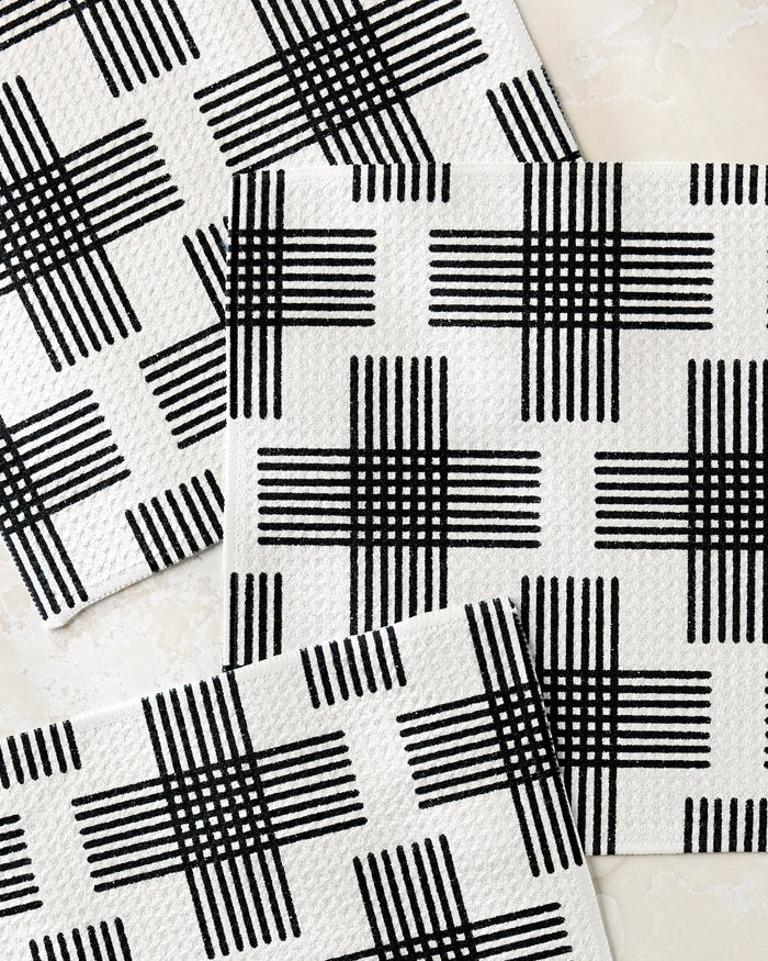 The Rosa Kichline Cross Hatch dishcloth set includes three white cloths with a black crisscross pattern, arranged overlapping at the corners. Made from recycled materials, they feature a modern geometric design perfect for any kitchen.