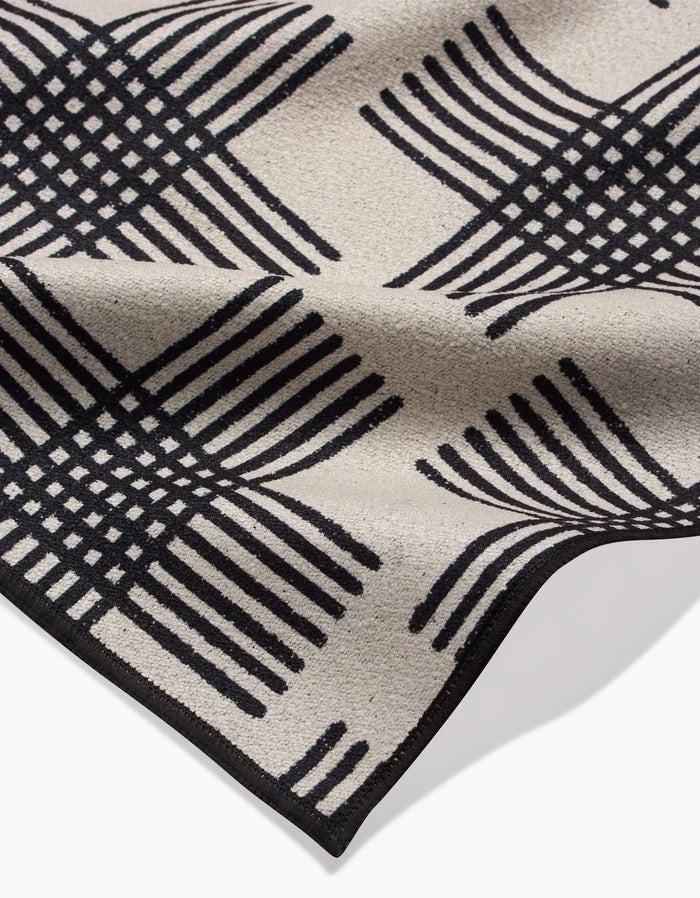 The Cross Hatch Beach Blanket by Rosa Kichline features a beige recycled fabric with a black geometric intersecting line design forming curved squares, shown slightly folded to highlight its textured surface and edge detail.