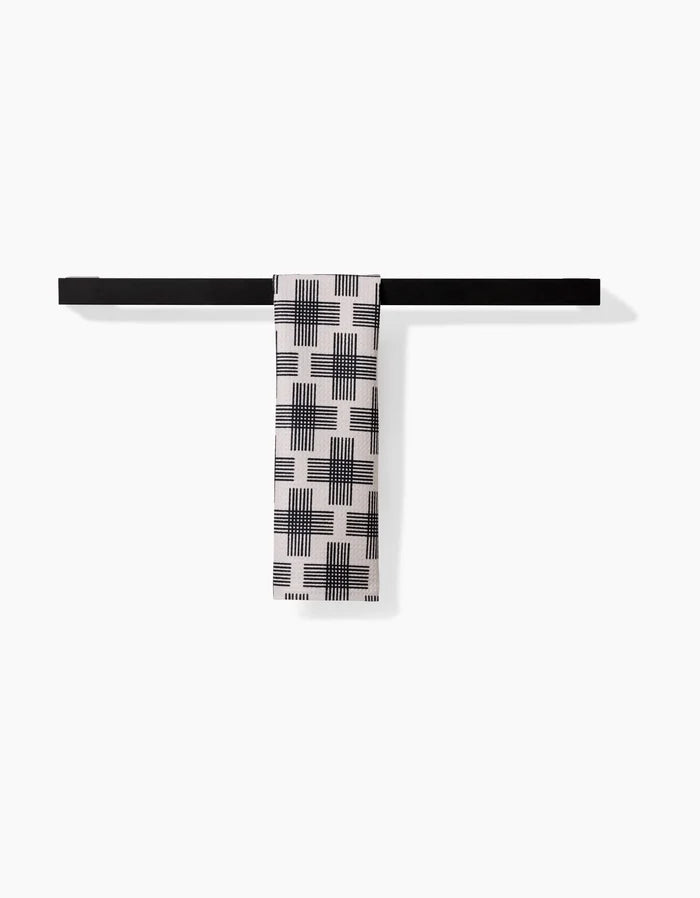 A black horizontal bar holds a folded white Bar Towel—Cross Hatch by Rosa Kichline. Made of Geoweave fabric, the towel displays a black geometric grid and line pattern against a plain white background.