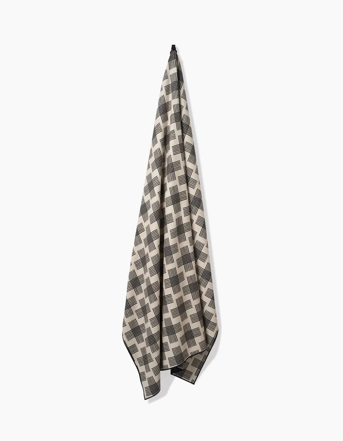 The Rosa Kichline Cross Hatch towel, made from recycled materials, hangs from a single hook against a white background. It features a beige and black geometric square and stripe design for an eco-friendly, stylish touch.