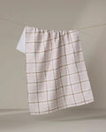 The Studio Jeannot Crossover is a white tea towel with a light brown grid, hanging over a clothesline against a beige background. Made from ultra-absorbent, recycled materials, it’s stylish and eco-friendly.