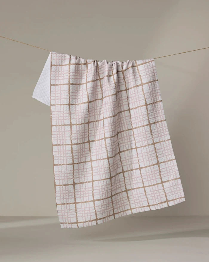 The Studio Jeannot Crossover is a white tea towel with a light brown grid, hanging over a clothesline against a beige background. Made from ultra-absorbent, recycled materials, it’s stylish and eco-friendly.