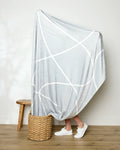 A person stands in a woven basket, mostly wrapped in Geometry's Crossover throw—a light gray blanket with white abstract lines. Only their lower legs and white sneakers show. A wooden stool rests nearby on the wood floor.