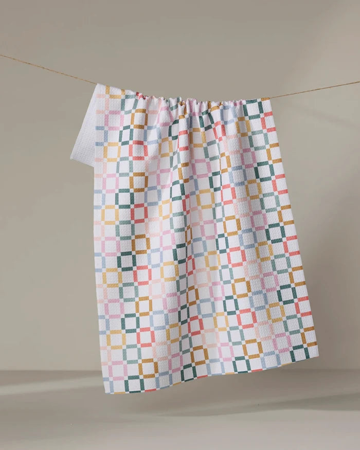 The "Crossroads" kitchen towel by Charlie Rowan Designs, featuring a colorful geometric square and grid pattern, hangs from a line against a plain beige background.