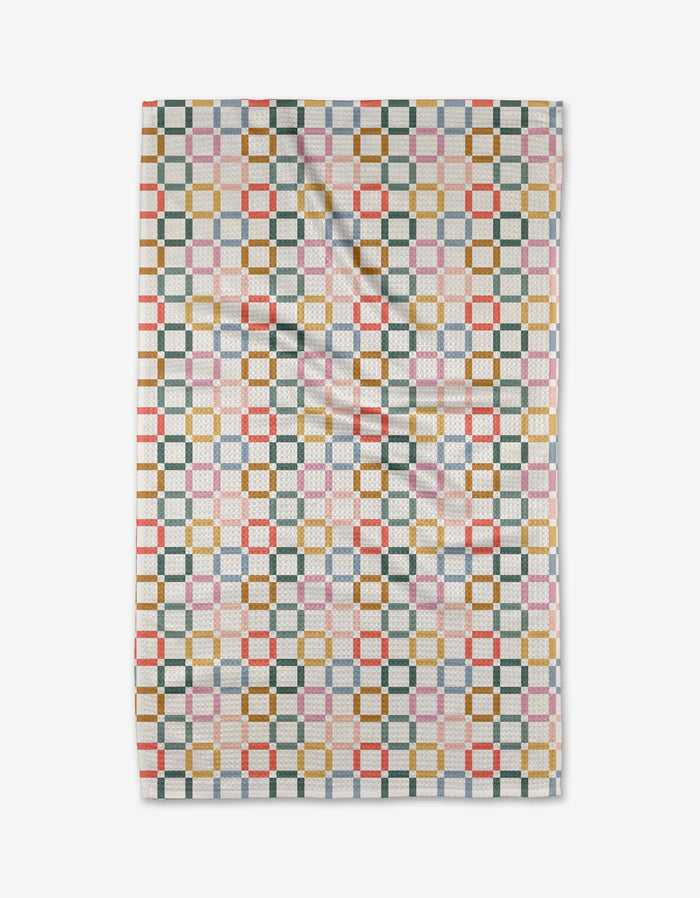 The Crossroads tea towel by Charlie Rowan Designs features a geometric pattern of interlocking squares in green, gold, red, blue, and pink on white Geoweave fabric, shown flat with slight folds.
