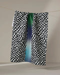 The Geometry "Current" tea towel, featuring a black-and-white checkered pattern with a green, blue, and purple gradient stripe, is made from ultra-absorbent recycled materials and hangs draped over a clothesline against a neutral backdrop.