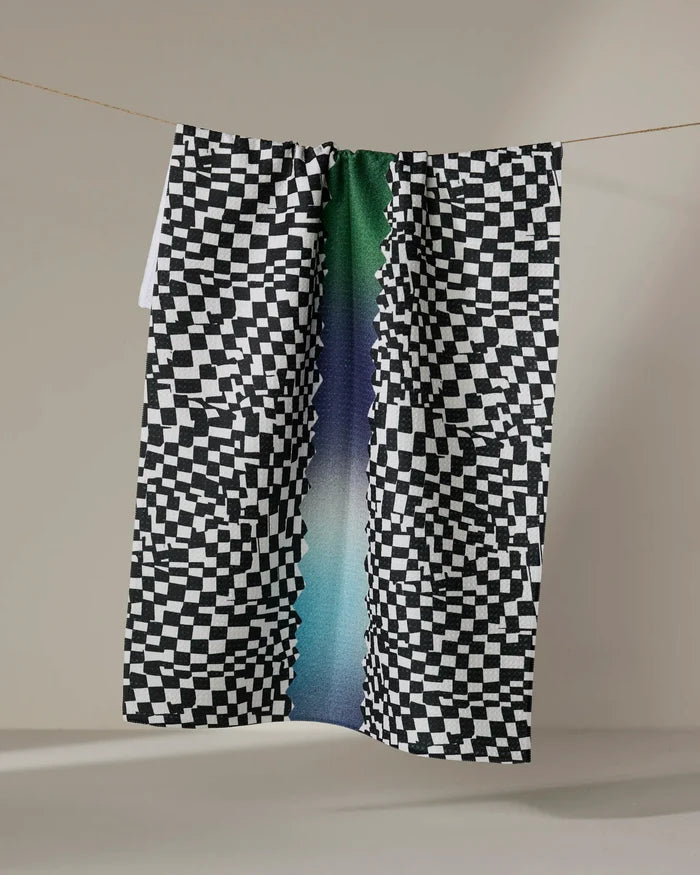 The Geometry "Current" tea towel, featuring a black-and-white checkered pattern with a green, blue, and purple gradient stripe, is made from ultra-absorbent recycled materials and hangs draped over a clothesline against a neutral backdrop.