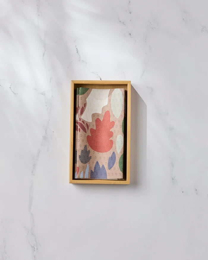 The Geometry Cut Paper Leaves Mocha notebook, featuring a colorful abstract leaf design made from post-consumer recycled materials, rests in a wooden tray on white marble, bathed in soft natural light.