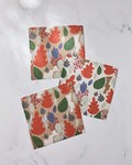 Three super-absorbent Geometry Cut Paper Leaves dishcloths, featuring colorful leaf and plant patterns in red, green, purple, and beige, are arranged on a white marble surface.