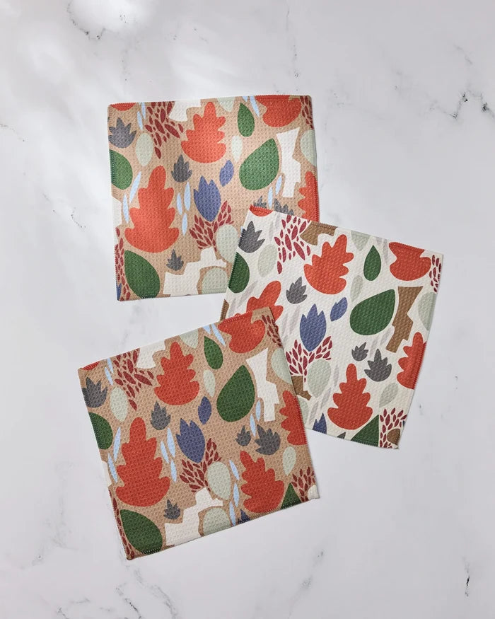 Three super-absorbent Geometry Cut Paper Leaves dishcloths, featuring colorful leaf and plant patterns in red, green, purple, and beige, are arranged on a white marble surface.