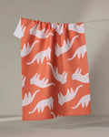 The Cute Cheetah tea towel by Jennifer Bouron features a coral color with white, stylized walking cats and colorful dots. Made from recycled materials, it hangs on a string against a neutral background.
