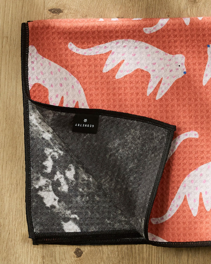 The Cute Cheetah by Jennifer Bouron is a coral blanket with white cat illustrations, pink spots, black trim, and a black brand tag. The corner is flipped to reveal the gray fleece underside. It rests on a wooden surface.