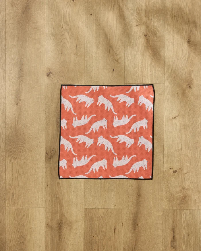 A rectangular orange ultra-absorbent "Cute Cheetah" dog towel by Jennifer Bouron, featuring white stylized cheetah silhouettes and a black border, lies on a light, natural-finish wooden floor.