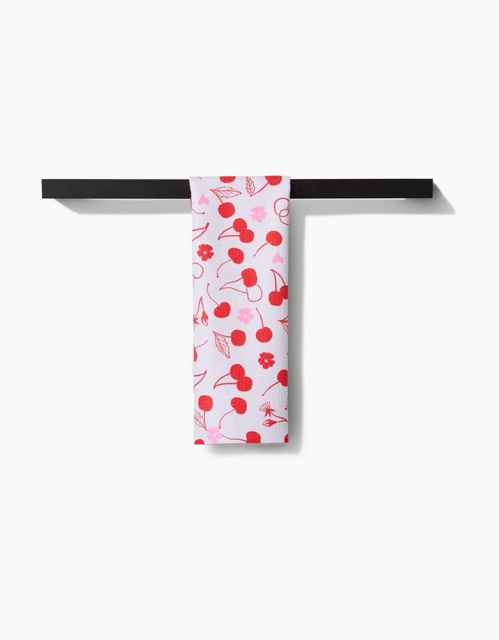 The Elana Gabrielle Cute Cherry white recycled kitchen towel, featuring cherry and flower patterns, hangs neatly over a horizontal black bar against a plain white background.