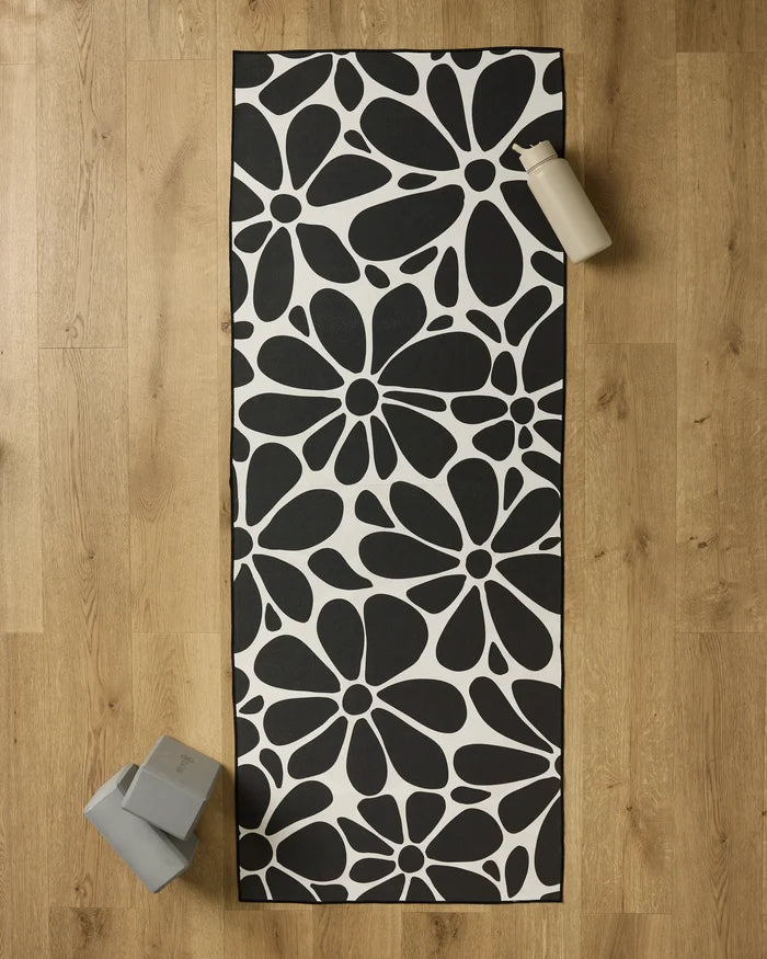 The GEOMETRY Cute Flowers yoga mat with a bold black and white floral pattern is laid on a wooden floor, topped with a non-slip towel. A water bottle and two yoga blocks sit near the mat’s corners.