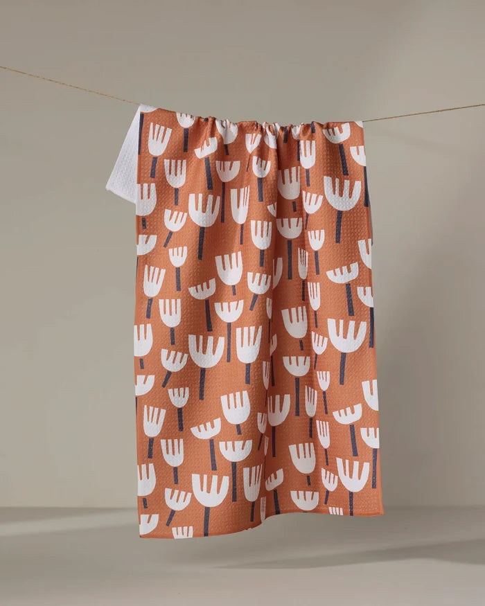 The "Cutout Flowers" tea towel by Claire Iglesias, featuring white tulip-shaped blooms with dark stems on an orange background, ultra-absorbent and made from recycled materials, hangs from a string against a neutral wall.
