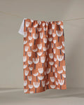 The "Cutout Flowers" tea towel by Claire Iglesias, featuring white tulip-shaped blooms with dark stems on an orange background, ultra-absorbent and made from recycled materials, hangs from a string against a neutral wall.