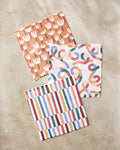The Claire Iglesias Cutout Shapes set includes three vibrant dishcloths—an orange and white floral, a multicolored swirl, and vertical stripes—all crafted from recycled materials on a beige surface.