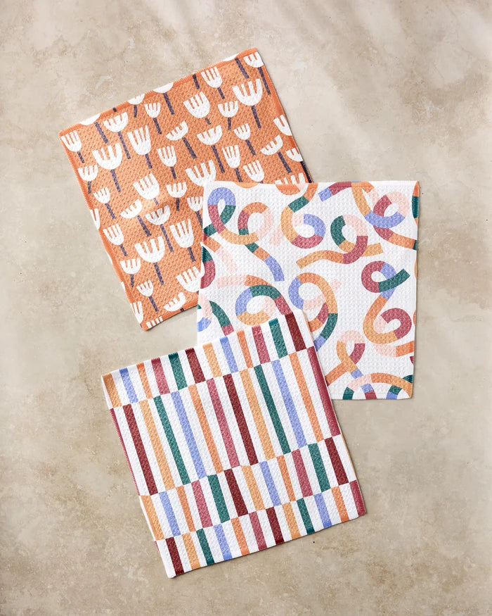 The Claire Iglesias Cutout Shapes set includes three vibrant dishcloths—an orange and white floral, a multicolored swirl, and vertical stripes—all crafted from recycled materials on a beige surface.