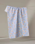 The Daisy Duets tea towel by Sarah Reed features orange polka dots and light blue abstract lines on white, made from recycled materials, and hangs on a clothesline against a neutral beige background.