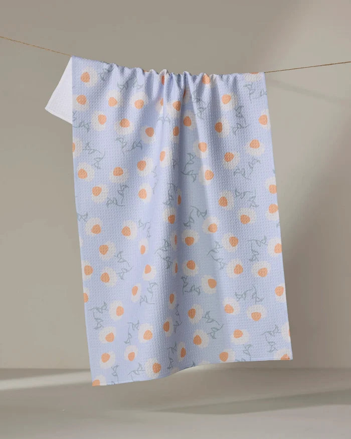 The Daisy Duets tea towel by Sarah Reed features orange polka dots and light blue abstract lines on white, made from recycled materials, and hangs on a clothesline against a neutral beige background.