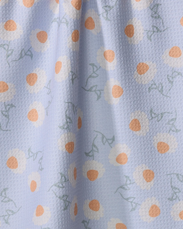 The "Daisy Duets" tea towel by Sarah Reed is light blue with a textured finish and features white and orange daisy-like flowers with light green stems, all made from recycled materials.