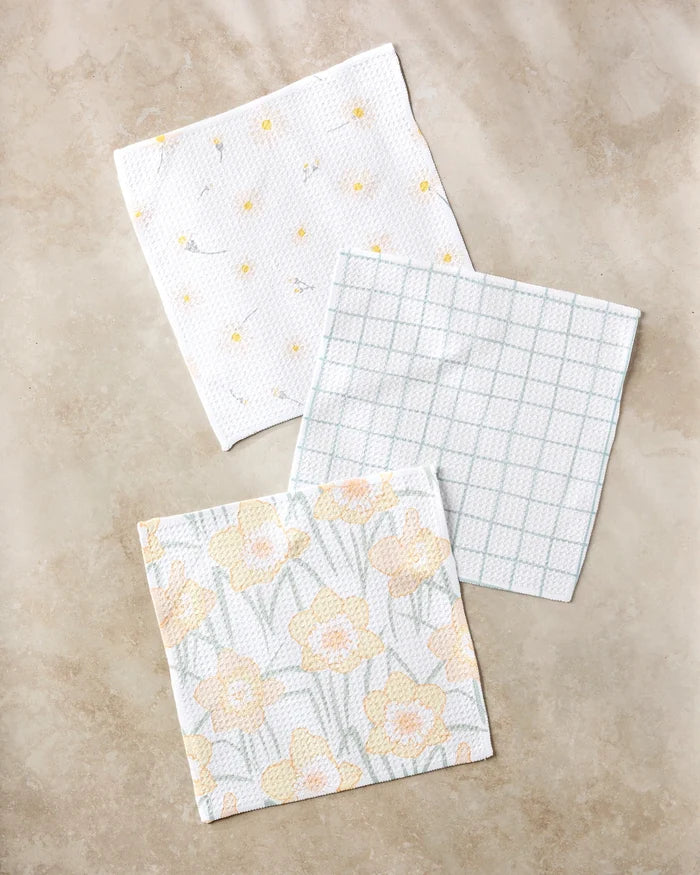 Three "Daisy Dust" patterned cloth napkins by Hufton Studio, made from recycled materials, are arranged on a beige surface—one with small yellow flowers, one in a blue grid pattern, and one with large yellow daffodils.
