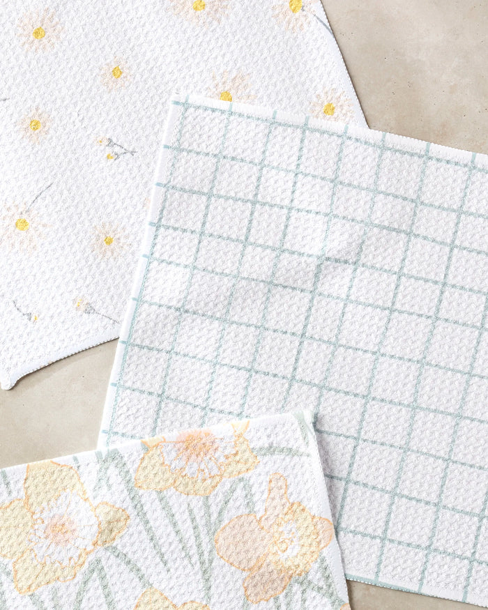 Three textured cleaning cloths from Hufton Studio’s Daisy Dust set, made from recycled materials, are displayed: one with a yellow floral pattern, one with a blue grid design, and one with yellow and gray spots.