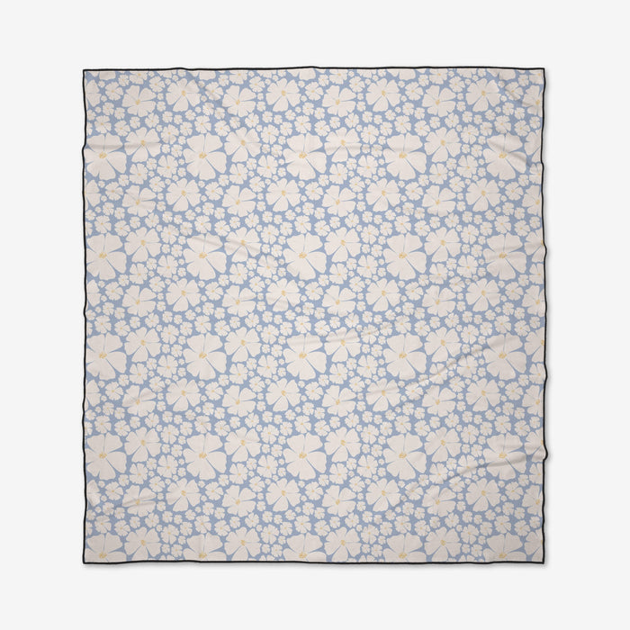 The Daisy Girl Blue beach blanket from Charlie Rowan Designs features a light blue background with white and light yellow hibiscus flowers with orange centers. Quick-drying and made from recycled materials, it’s finished with a thin dark border.