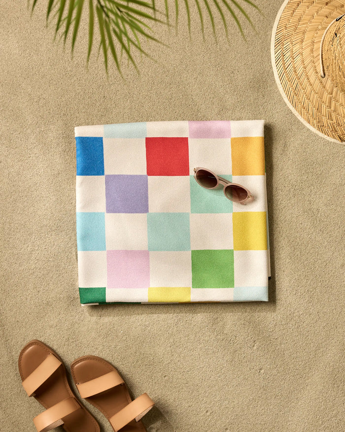 A folded "Dance Floor" beach blanket by Vaishali Design Studio with a colorful checkered pattern lies on the sand beside brown sandals, sunglasses, a woven hat, and a palm leaf casting its shadow.