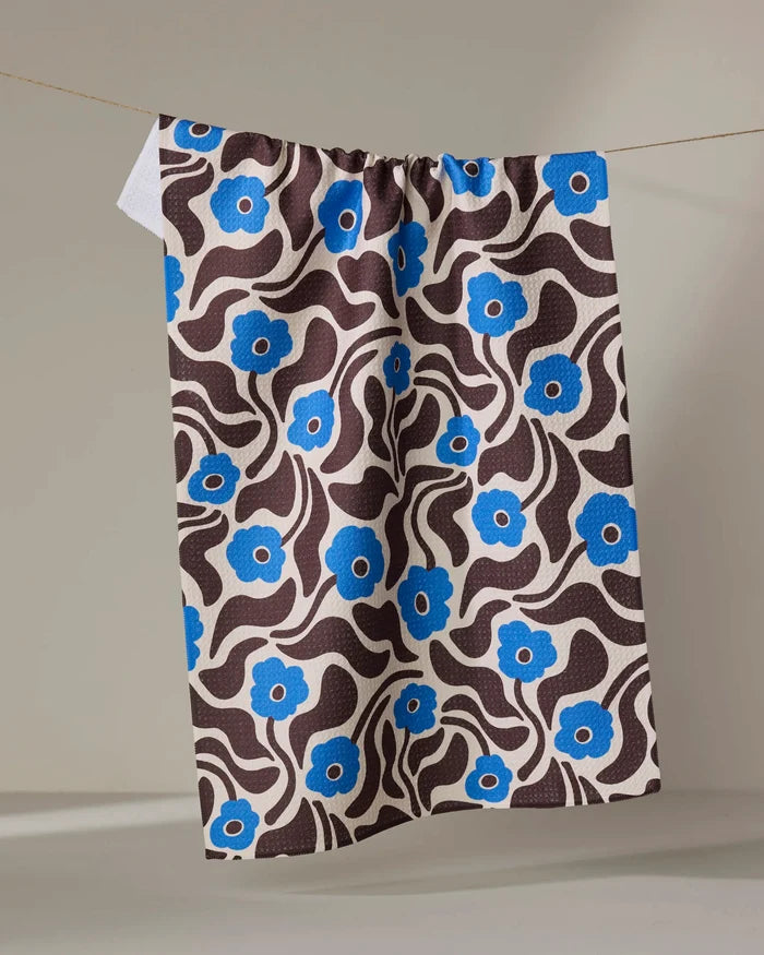 The Gerdadzy "Dancing Blooms" tea towel, made from recycled materials, features a brown and white wavy pattern with bright blue flowers and hangs from a string against a plain beige background.
