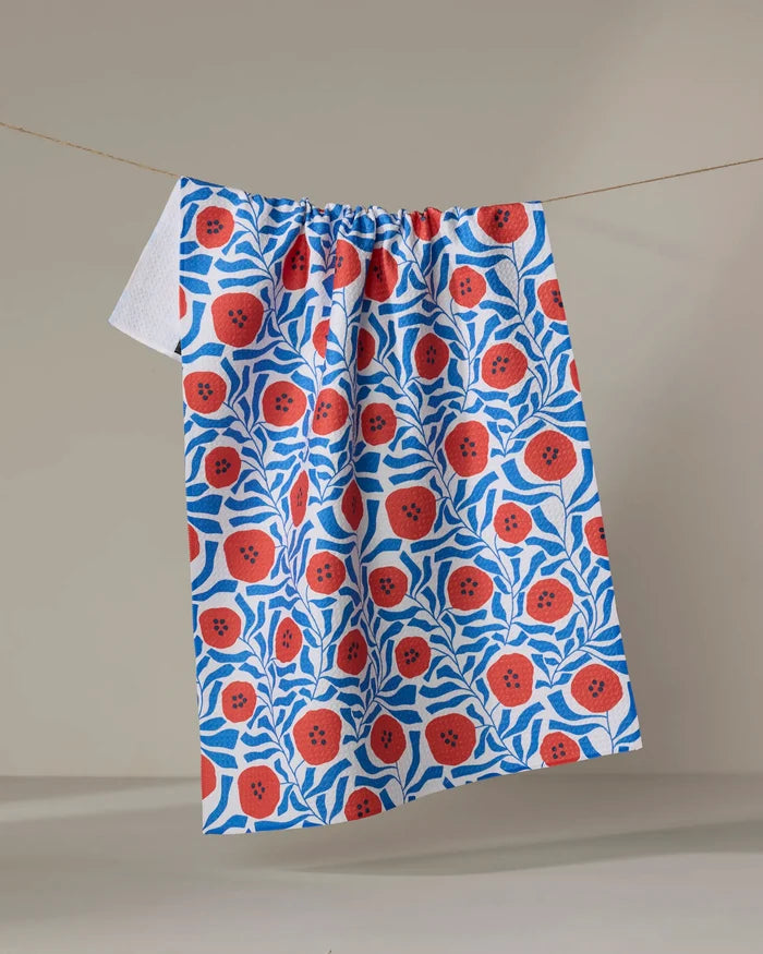 The Dancing Poppies tea towel by Happy Hemera, made from recycled materials, displays vibrant red flowers and blue leaves and hangs on a clothesline against a neutral background.