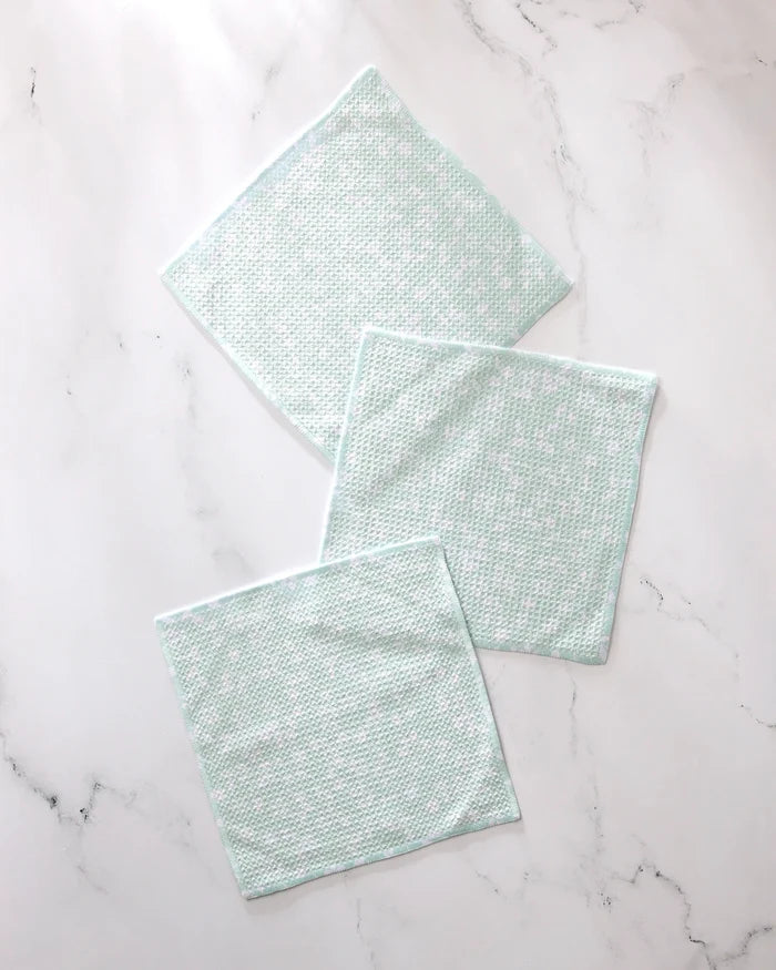 Three Dandy reusable dishcloths by GEOMETRY, featuring a light green and white textured design, are spread out on a white marble surface with their edges slightly overlapping.