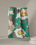 The Geometry Darla tea towel, made from quick-drying recycled materials, features bold abstract floral prints in white, yellow, green, and pink. It hangs on a line near stacked white plates against a neutral background.