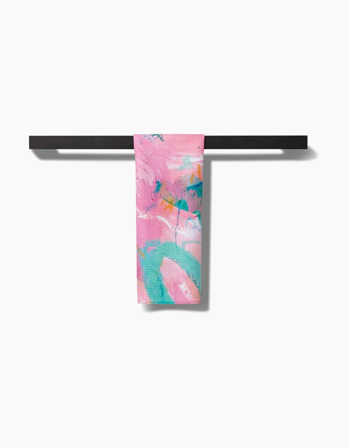The Daughter bar towel by Sarah Holden, made from soft Geoweave fabric for superior absorbency, features abstract turquoise, white, and coral patterns on pink and hangs neatly on a black towel bar against a white background.