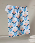 The Daytime Florals tea towel by Blissful Flamingo, crafted from recycled materials, features a blue, pink, and tan geometric floral design. It hangs on a line with a light background and stacked white dishes in the corner. Eco-friendly and ultra-absorbent.