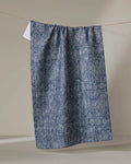 The Debra Blue tea towel by Becki Owens, crafted from recycled materials, features blue and white textures and hangs neatly from a clothesline against a neutral background.