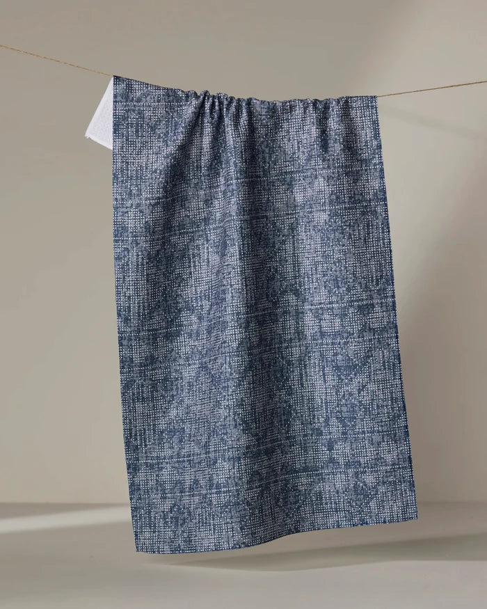 The Debra Blue tea towel by Becki Owens, crafted from recycled materials, features blue and white textures and hangs neatly from a clothesline against a neutral background.