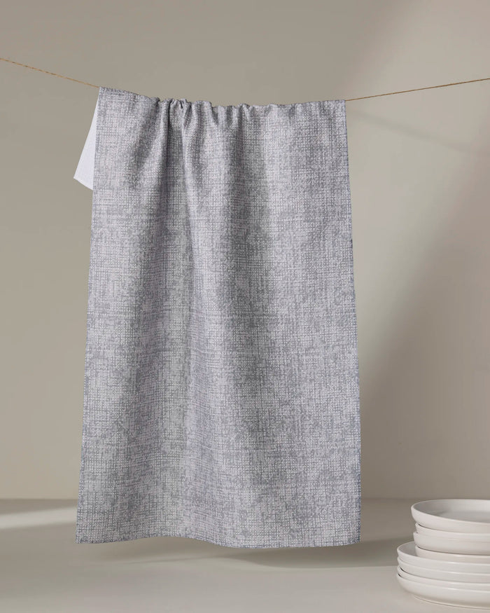The Debra Grey kitchen towel by Becki Owens hangs from a string against a neutral background, with a stack of white plates placed nearby.