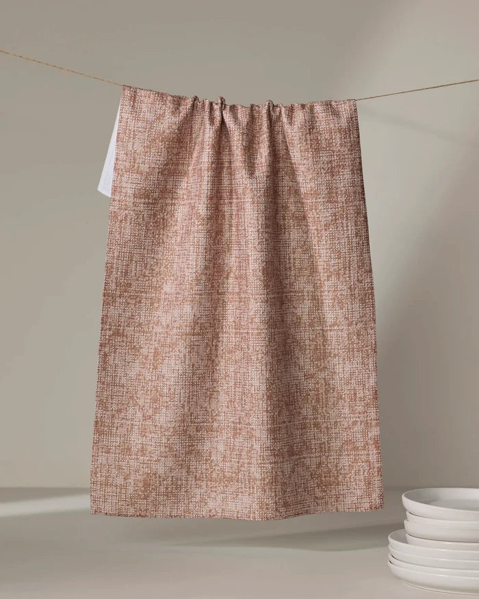 The Debra Rust recycled kitchen towel by Becki Owens hangs on a clothesline against a neutral background, with a stack of white plates nearby.