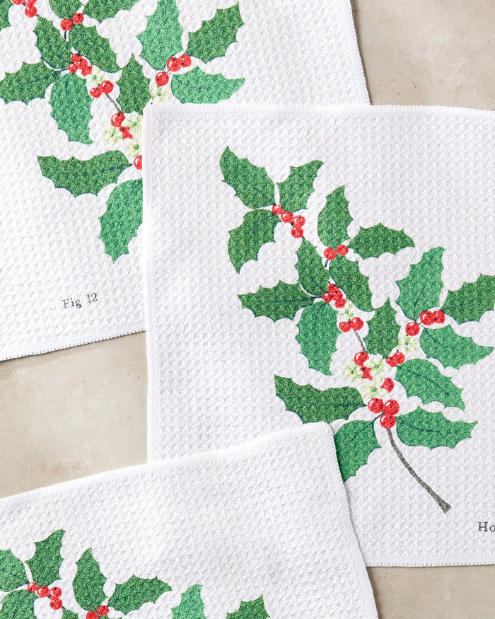 Three white "December Holly" dishcloths by Linda Fox, made from recycled materials, display green holly leaves, red berries, and script text on each. The botanical-style cloths are shown on a neutral background.