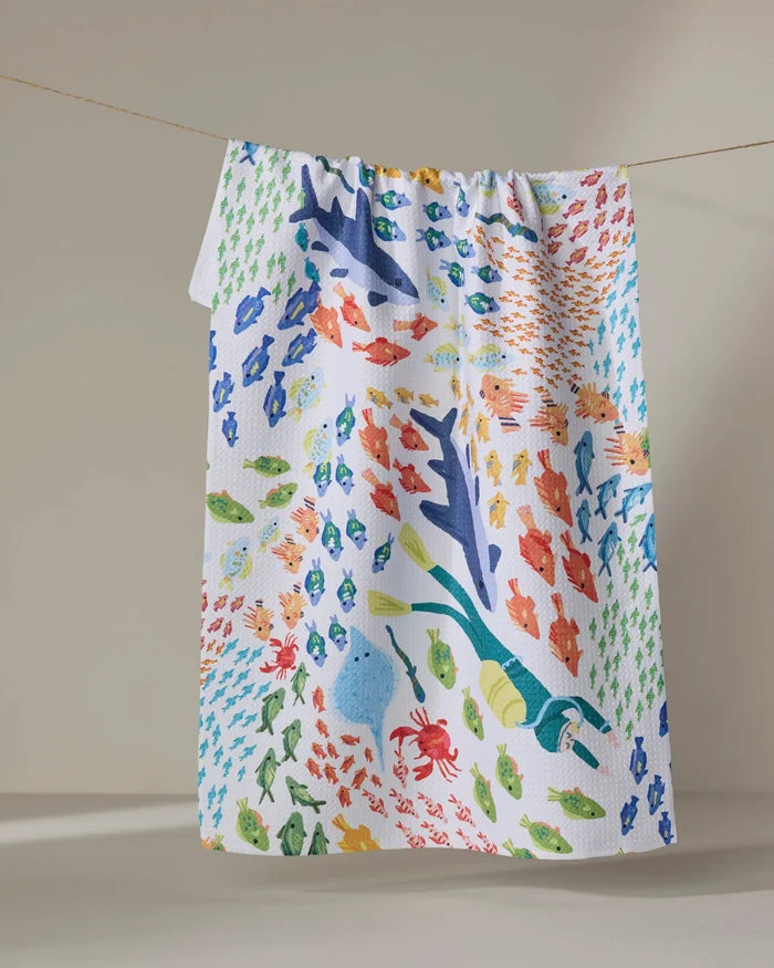 The "Deep Sea Encounter" tea towel by Janna Sue Design features vibrant sharks, fish, crabs, and seaweed, sustainably crafted from recycled materials and shown hanging on a clothesline against a simple backdrop.