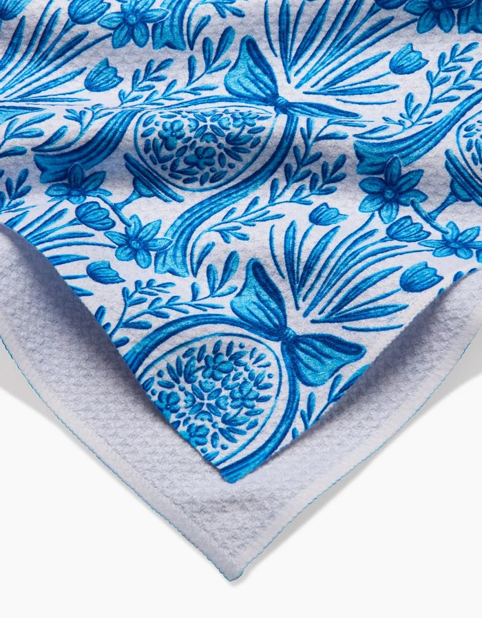 A close-up of the Sarah Daugherty Delft Blue Easter Egg Damask towel, ultra-absorbent and white, shows its blue floral and pomegranate pattern with leaves and flowers, folded to reveal both the front design and terry cloth underside.