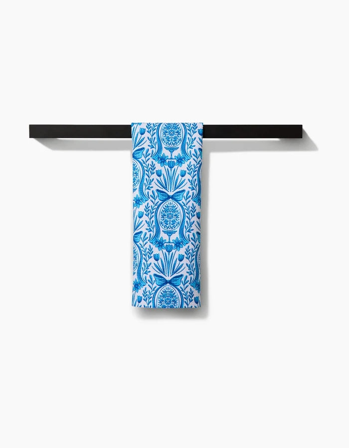 The Sarah Daugherty Delft Blue Easter Egg Damask tea towel, featuring a blue and white floral and tree pattern, hangs neatly over a simple black towel bar against a white background.
