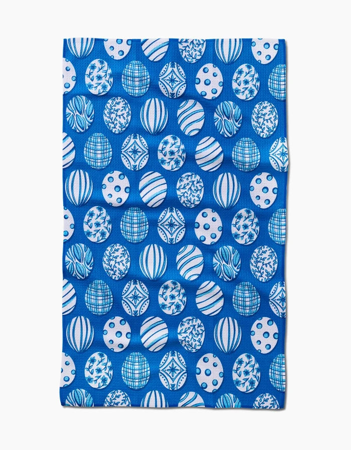 The Delft Blue Easter Eggs tea towel by Sarah Daugherty is rectangular, blue, and made from recycled materials, featuring a repeating pattern of white and blue spheres with geometric and abstract designs.