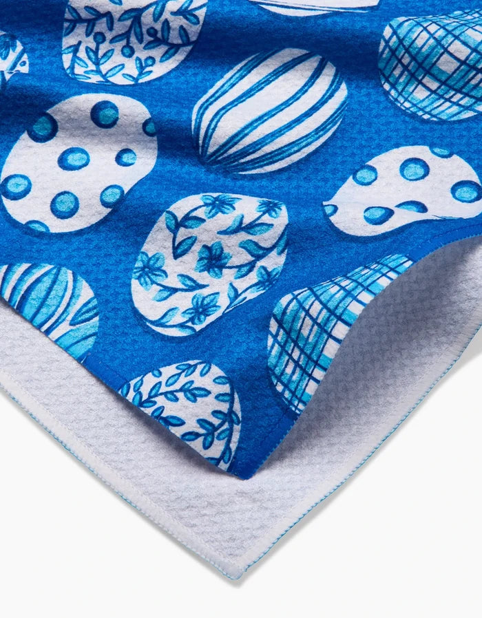 The Delft Blue Easter Eggs tea towel by Sarah Daugherty features blue and white ovals with floral, striped, and polka dot designs. Made from quick-drying recycled materials, it has a textured white underside and blue-stitched edging.