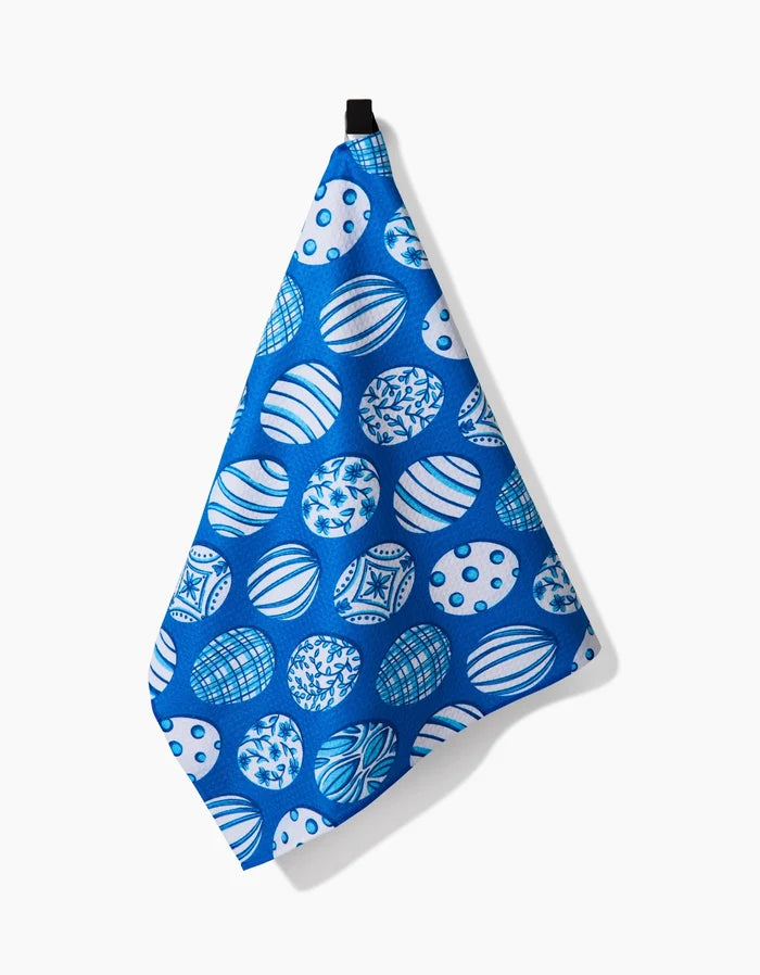 The “Delft Blue Easter Eggs” tea towel by Sarah Daugherty, featuring quick-drying fabric and a blue pattern of decorative spheres, hangs from a black loop against a white background.